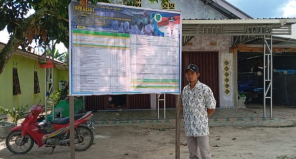 Kepala Desa Kartamulia, Aman, menunjukkan baliho APBDes di salah satu RT/RW sebagai bentuk transparansi pengelolaan keuangan desa.