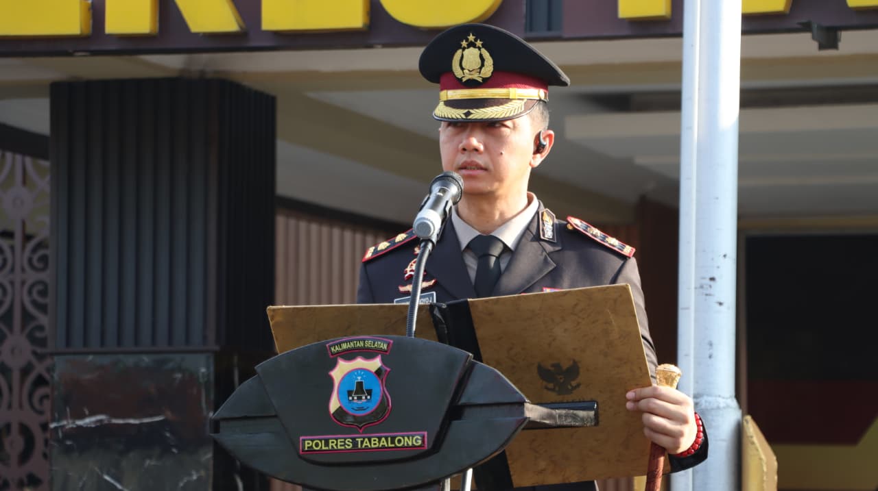 Polisi berpangkat di podium membacakan teks, peserta upacara baris rapi di belakang