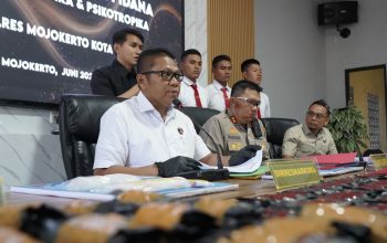Tiga polisi duduk di meja konferensi pers, latar spanduk biru Ditresnarkoba Polda Jatim