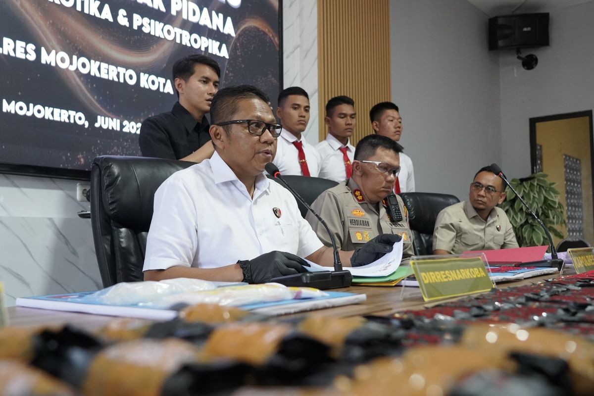 Tiga polisi duduk di meja konferensi pers, latar spanduk biru Ditresnarkoba Polda Jatim