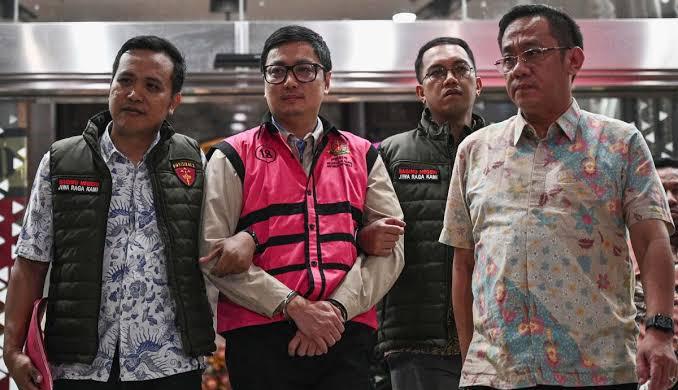 Empat pria berjas berjalan, satu di antaranya digiring ke mobil tahanan